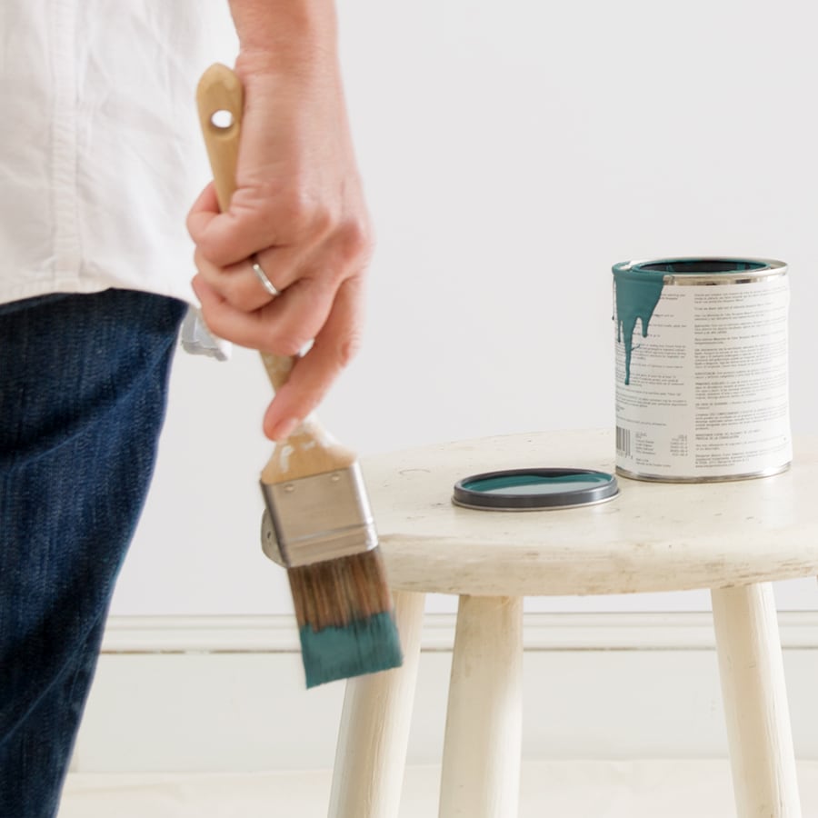 A woman's hand holding a paint brush with green paint on it, with an open Benjamin Moore paint color sample pint on the table next to her.