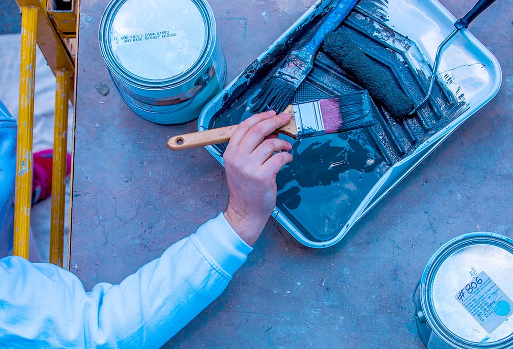 A contractor holding a paint brush after dipping in a paint tray with two gallons Benjamin Moore of paint.
