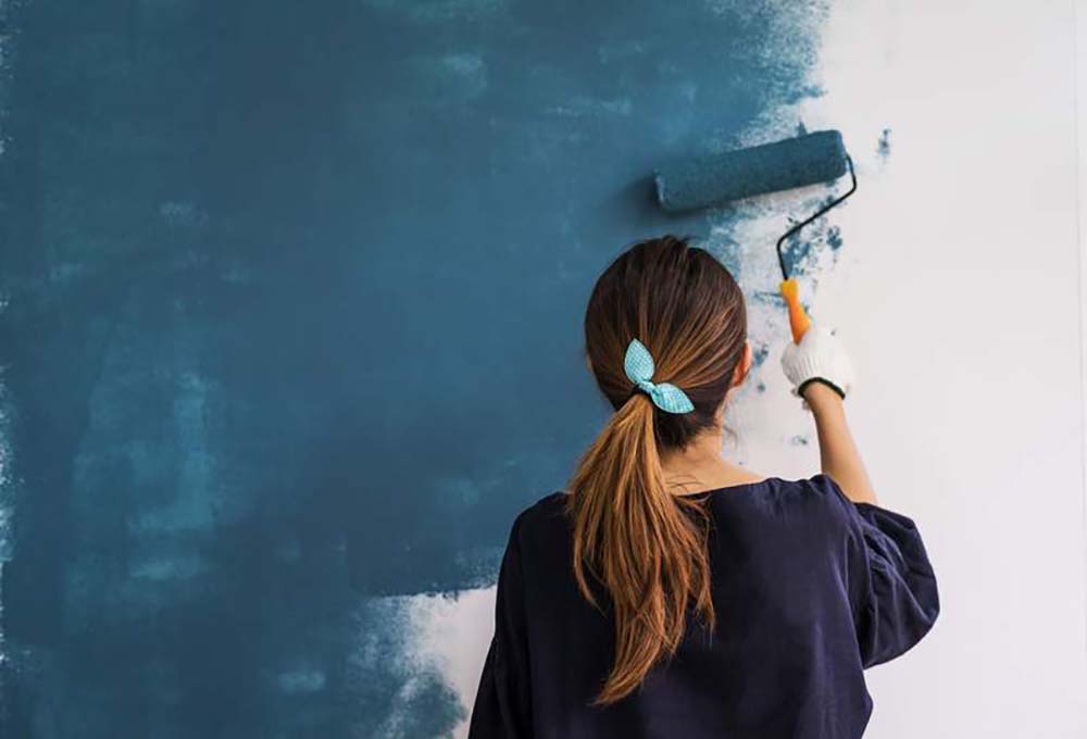 Back of a woman painting a wall dark blue with a paint roller.