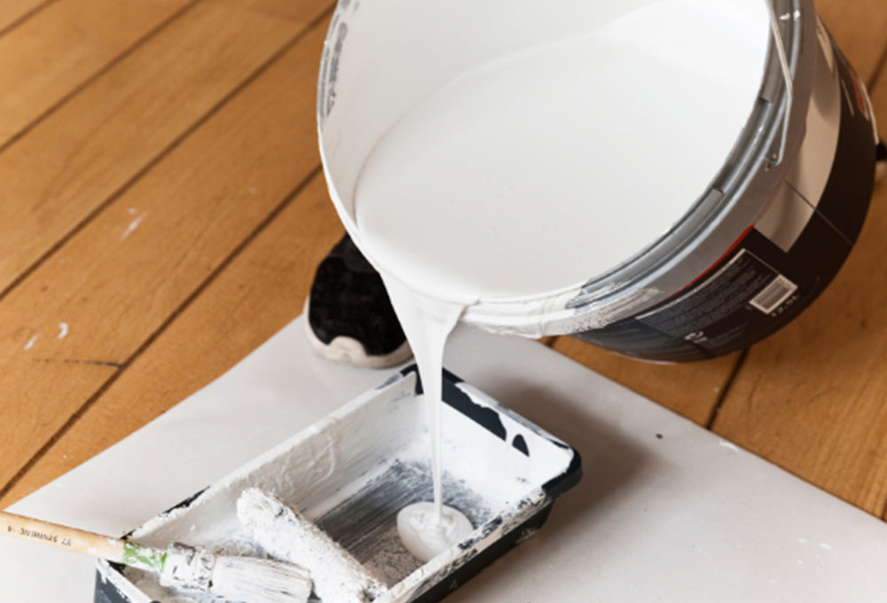 A 5 gallon pail of primer being poured into a paint tray with a paint roller and paint brush.