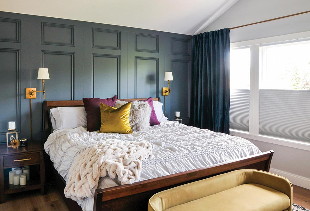 A bedroom with a dark accent wall, with a wood bed and gold and purple pillows with a white duvet.