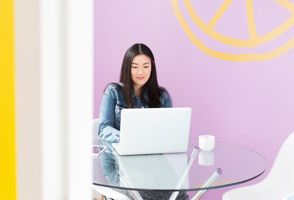 A woman sitting at a table in front of a laptop in front of a purple and yellow wall. 