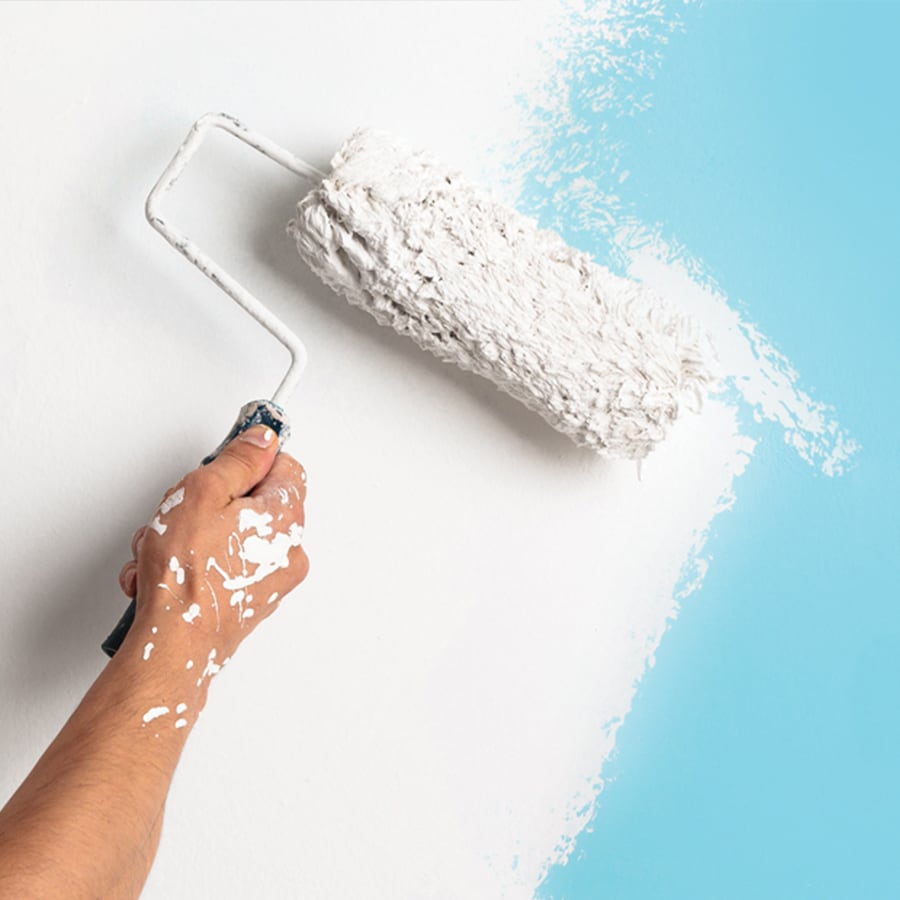 A person's hand with a paint roller, rolling white paint onto a light blue wall.