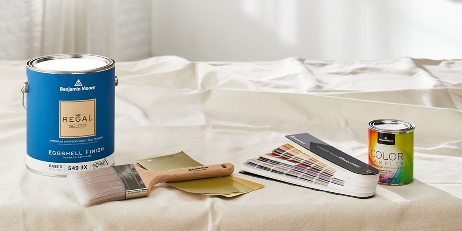 Benjamin Moore eggshell finish paint can, color samples paint can, a paint brush, and a fan deck all set on top of a white tarp.